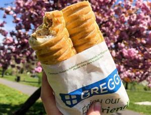 Someone holding up a Greggs vegan sausage roll with a flowering tree in the background
