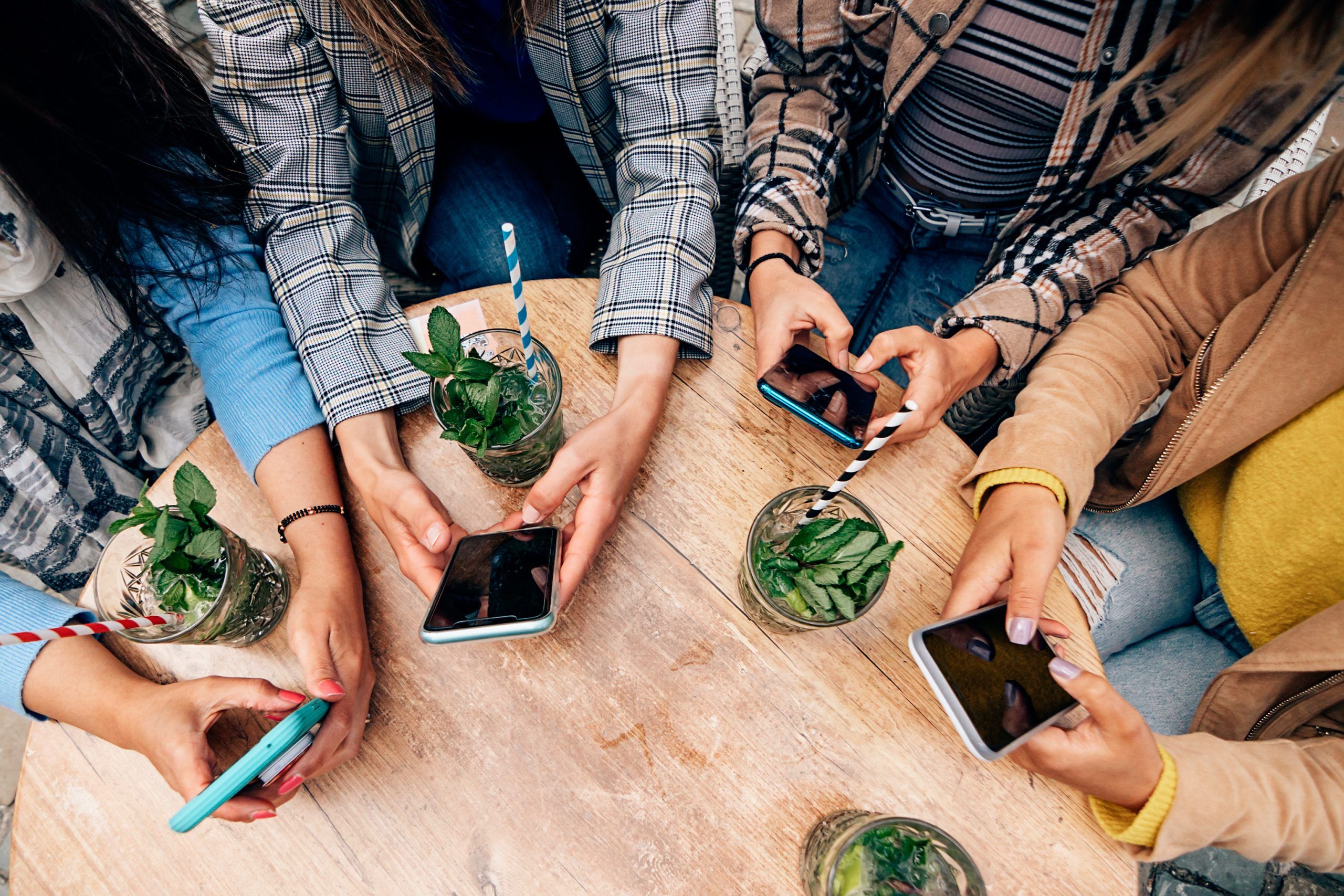 Four women sitting around a wooden table with drinks, all holding smartphones