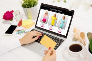 A woman at her desk with her laptop open to an online fashion shop. She's choosing between a pink, yellow, and light blue dress. Around here are several items such as a potted plant, a seashell, her phone, and a cup of coffee.