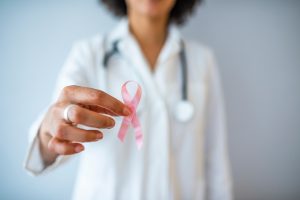 A female doctor stands in front of a while wall. Her body is out of focus, but she's wearing a white coat and a stethoscope. The focus is on the hand she holds out, which holds a pink ribbon as a symbol for the fight against breast cancer.