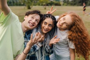 Three children are grouped up and taking a selfie. To the left is a boy with brown hair and a green shirt. In the middle is a girl of colour with braids and a blue plaid shirt. At the right is a redhead girl in a white shirt. They're in a field and all smiling.